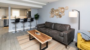 A modern living room with a grey sofa, wooden coffee table, and a yellow armchair. A kitchen with bar stools is visible in the background. - A Private Boho-Modern Stay Minutes to Downtown w/ KING Bed and Hidden Nook! (Des Moines)