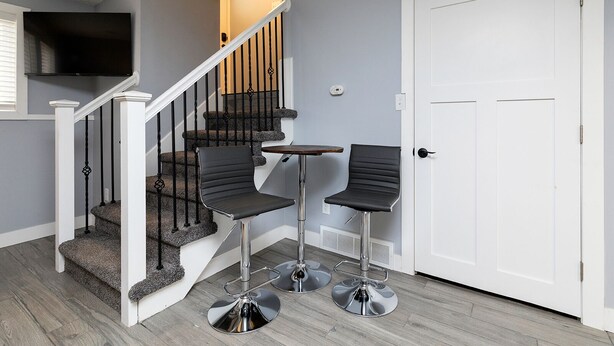 A staircase with a TV mounted above, two bar stools, and a white door. - A Private Boho-Modern Stay Minutes to Downtown w/ KING Bed and Hidden Nook! (Des Moines)