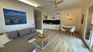 A modern living room with a grey sofa, a wooden coffee table, and a painting of a coastal scene. A kitchen with white cabinets and a dining area with a white table and chairs is visible in the background. - Casa di Alessia THE new vacation apartment with dream view over Malcesine incl. pool (Malcesine)