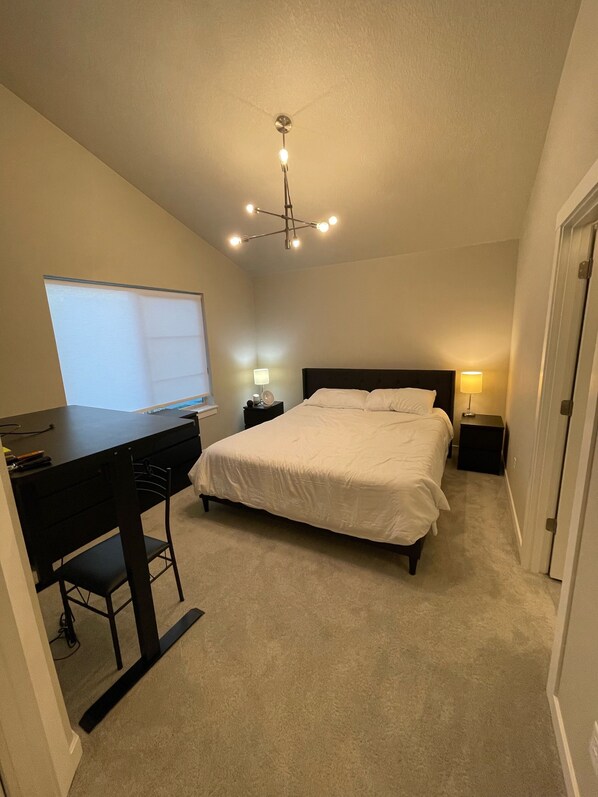 A bedroom with a bed, a desk, and a chair. There is a window, a ceiling light, and a door.
