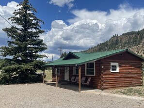 A rustic log cabin with a green roof, a porch with chairs, and a view of a mountainous landscape.