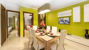 Ein moderner Essbereich mit einem Holztisch, gedeckt für vier Personen, umgeben von weißen Stühlen. Eine große Flachbildfernseher ist rechts an der Wand befestigt.