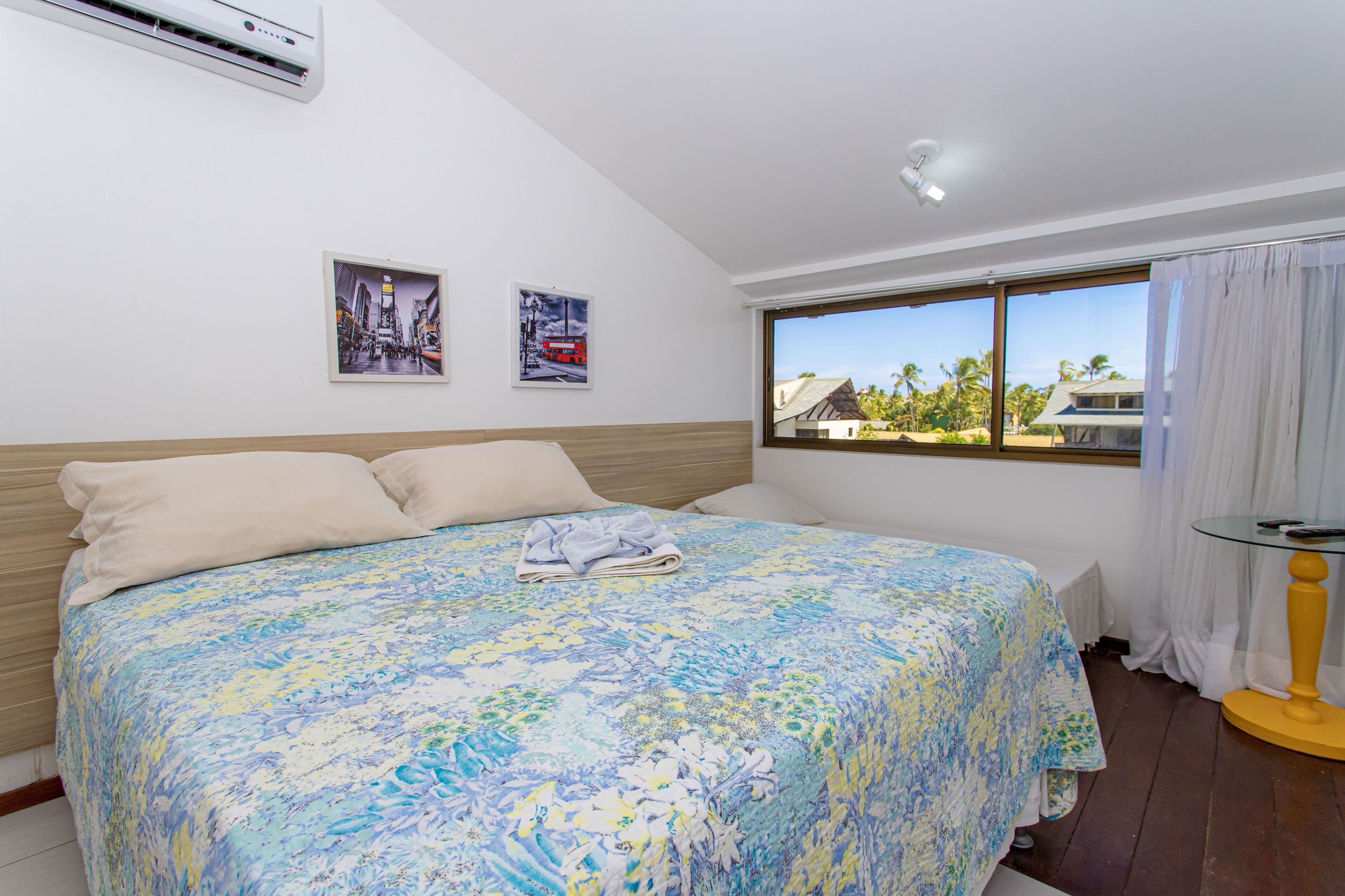 A bedroom with a large bed, a window with a view of buildings and palm trees, and a small table with a lamp.