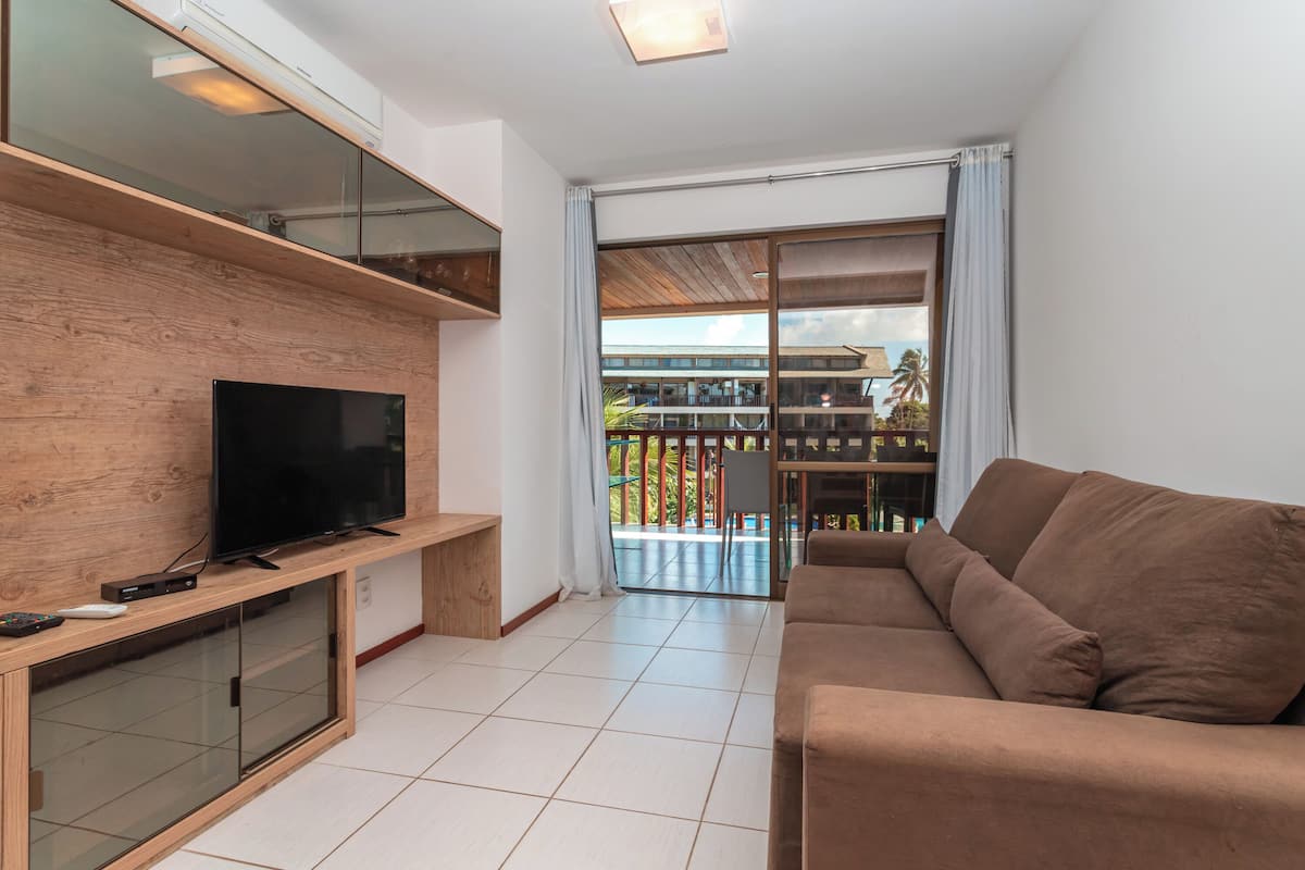 Sala moderna com televisão de tela plana sobre console de madeira, sofá marrom e porta de vidro que dá para uma varanda com vista para um edifício e palmeiras.