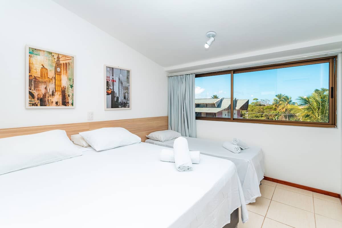 Quarto de hotel com duas camas, uma janela grande com vista para palmeiras e duas obras de arte penduradas na parede.