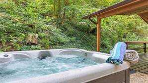 A hot tub with a wooden deck and a wooden pergola, surrounded by lush greenery.