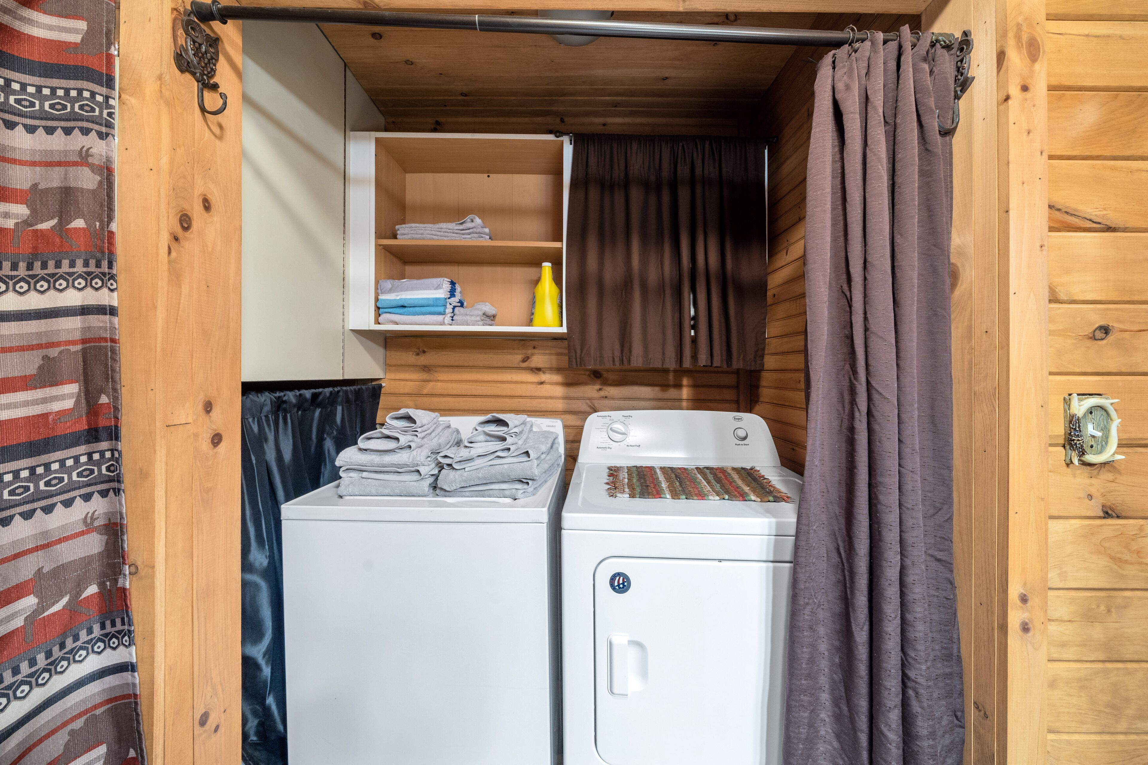 A compact laundry area with two washing machines, a shelf with folded towels, and a curtain.
