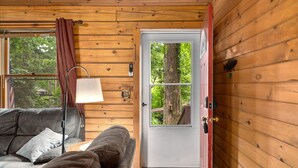 A cozy room with a wooden interior, a leather sofa, a lamp, and a door leading to an outdoor area with a tree.