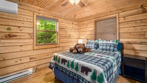 A wooden cabin bedroom with a bed, a teddy bear, a ceiling fan, and a window with a view of greenery.