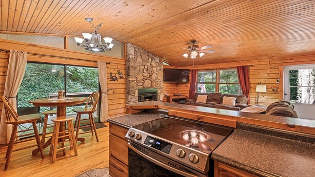 A cozy cabin interior with a stone fireplace, wooden dining area, kitchen with a stove, and a living area with a couch and TV.