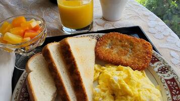 Un plato de desayuno con tostadas, salchicha, huevos revueltos y un alimento frito, acompañado de un vaso de jugo de naranja y una taza de café, sobre una mesa con vista a un jardín y casas.