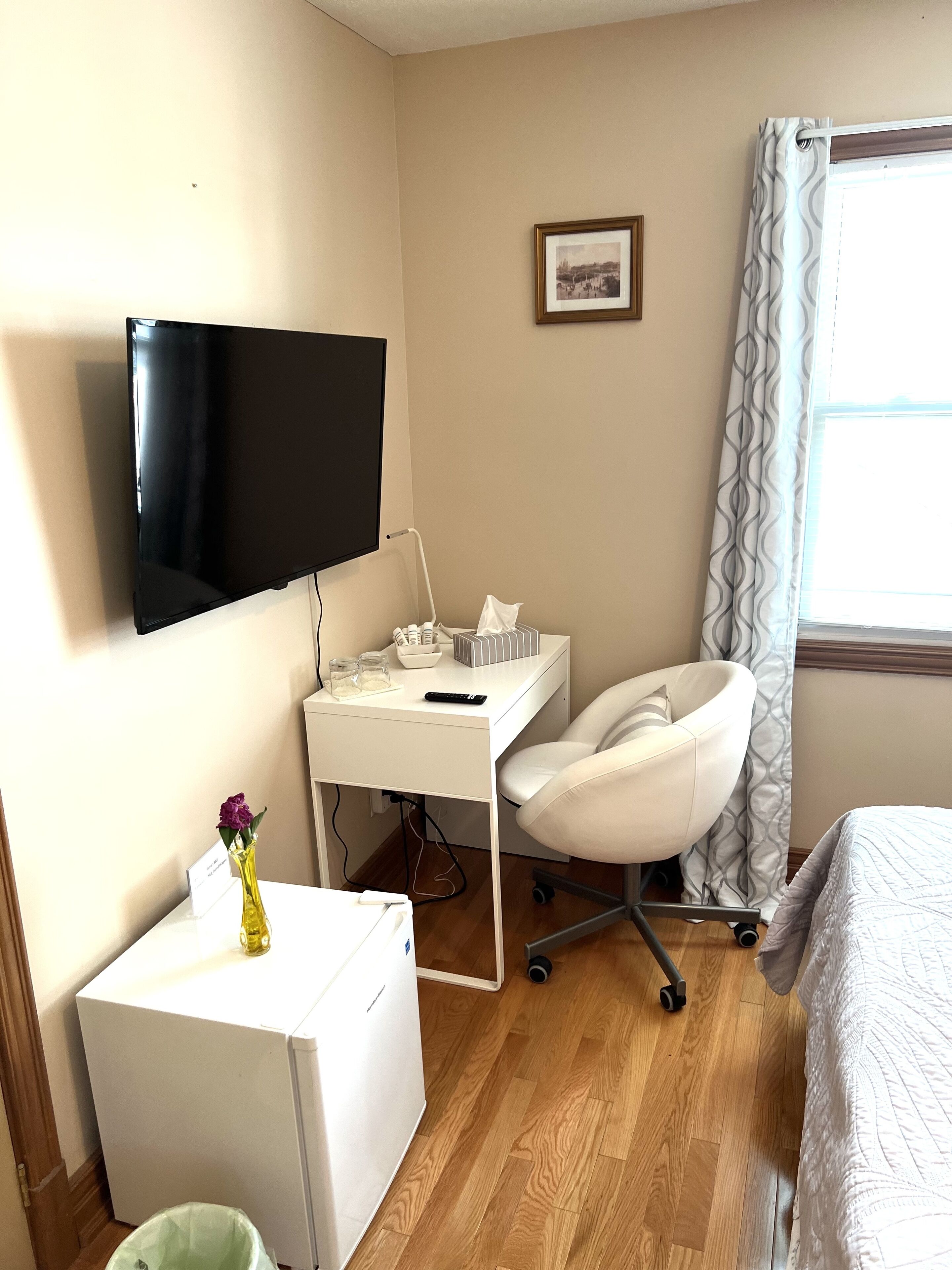 Habitación de hotel con televisor de pantalla plana, escritorio con silla, refrigerador pequeño y cama con colcha blanca.