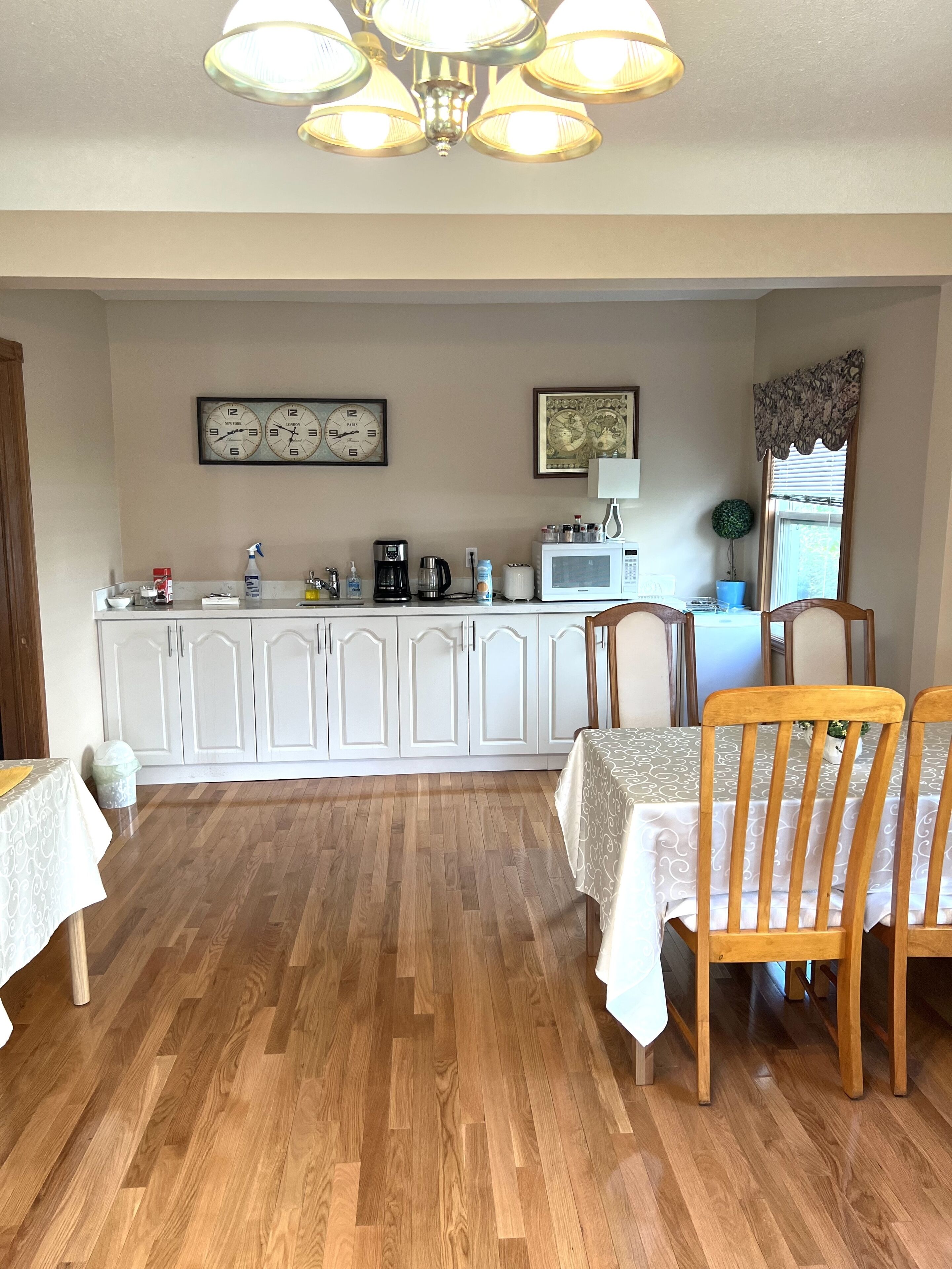 A dining area with wooden chairs and a table, a kitchen with white cabinets, a clock, and a framed picture on the wall.