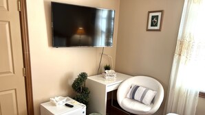 A hotel room with a bed, a desk with a chair, a television, a small refrigerator, a trash bin, and a window with curtains. - Anna's Bed & Breakfast (Niagara Falls)