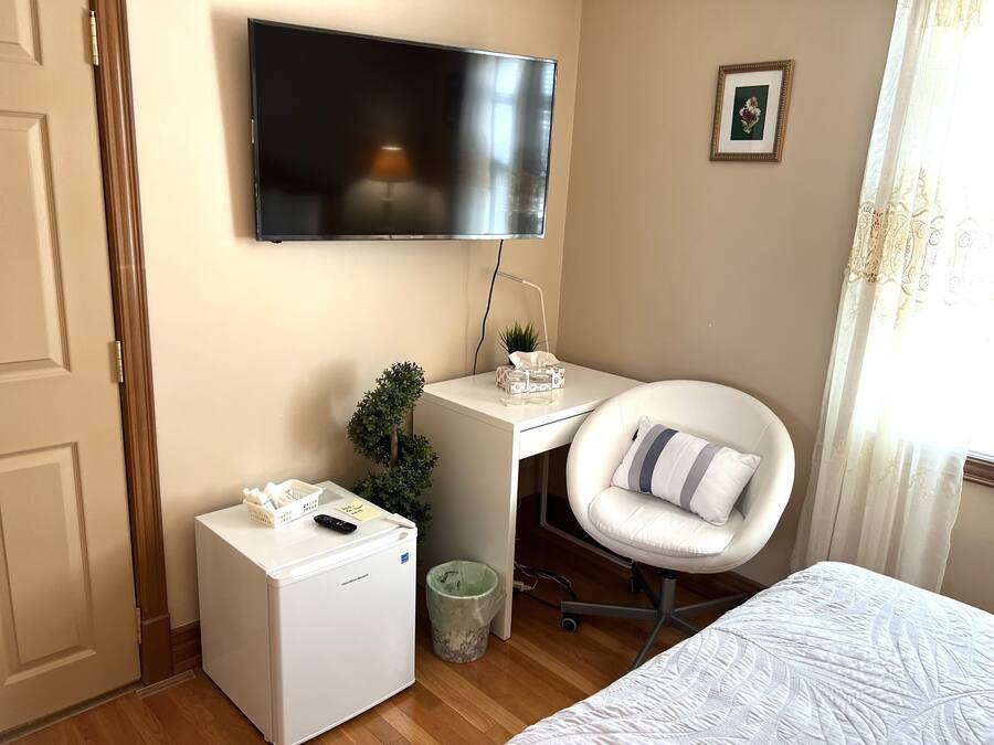 Quarto de hotel com cama, escrivaninha com cadeira, televisão, mini-frigorífico, lixeira e janela com cortinas.