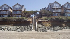 A staircase leading to a beachfront property with multiple balconies, a rocky shoreline, and a clear sky.