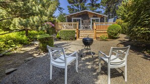 A cabin with a gravel patio, two white chairs, a grill, and a wooden deck.