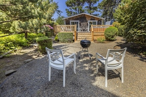 A cabin with a gravel patio, two white chairs, a grill, and a wooden deck.