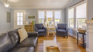 A living room with a leather sofa, two armchairs, a wooden coffee table, and a side table with a lamp. There are three windows with blinds, a door, and a sign that reads 'Home Store'.