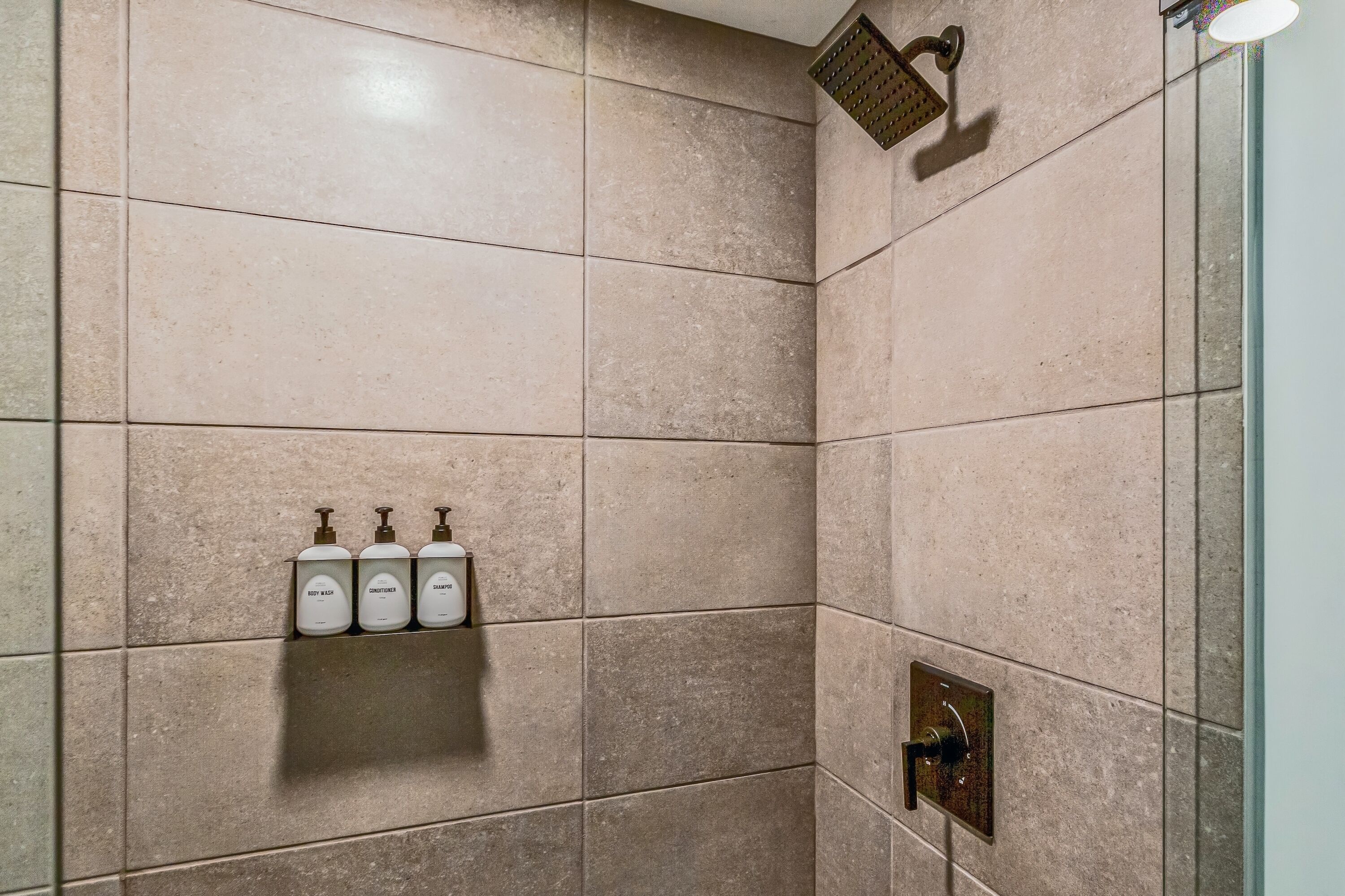 Et bruseområde med beige fliser, et brusehoved og tre toiletartikler på en hylde.