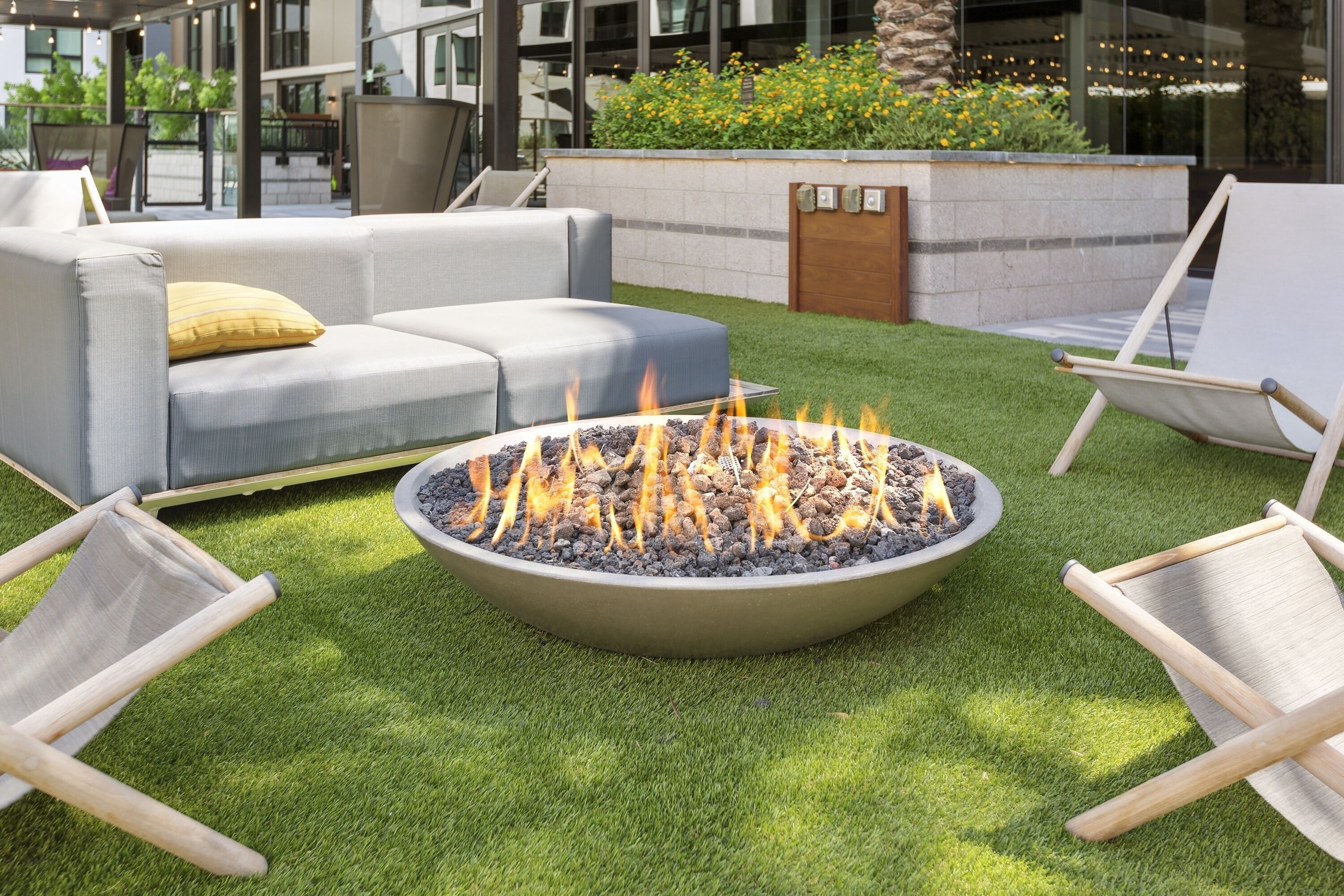 A fire pit surrounded by a grey sofa and two wooden chairs on a well-maintained lawn.
