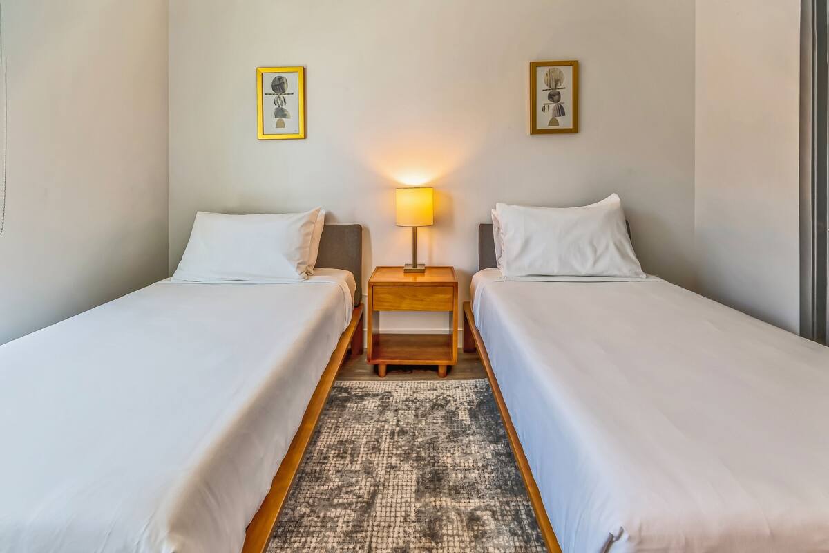 A hotel room with two single beds, a small wooden nightstand with a lamp, and a patterned rug.