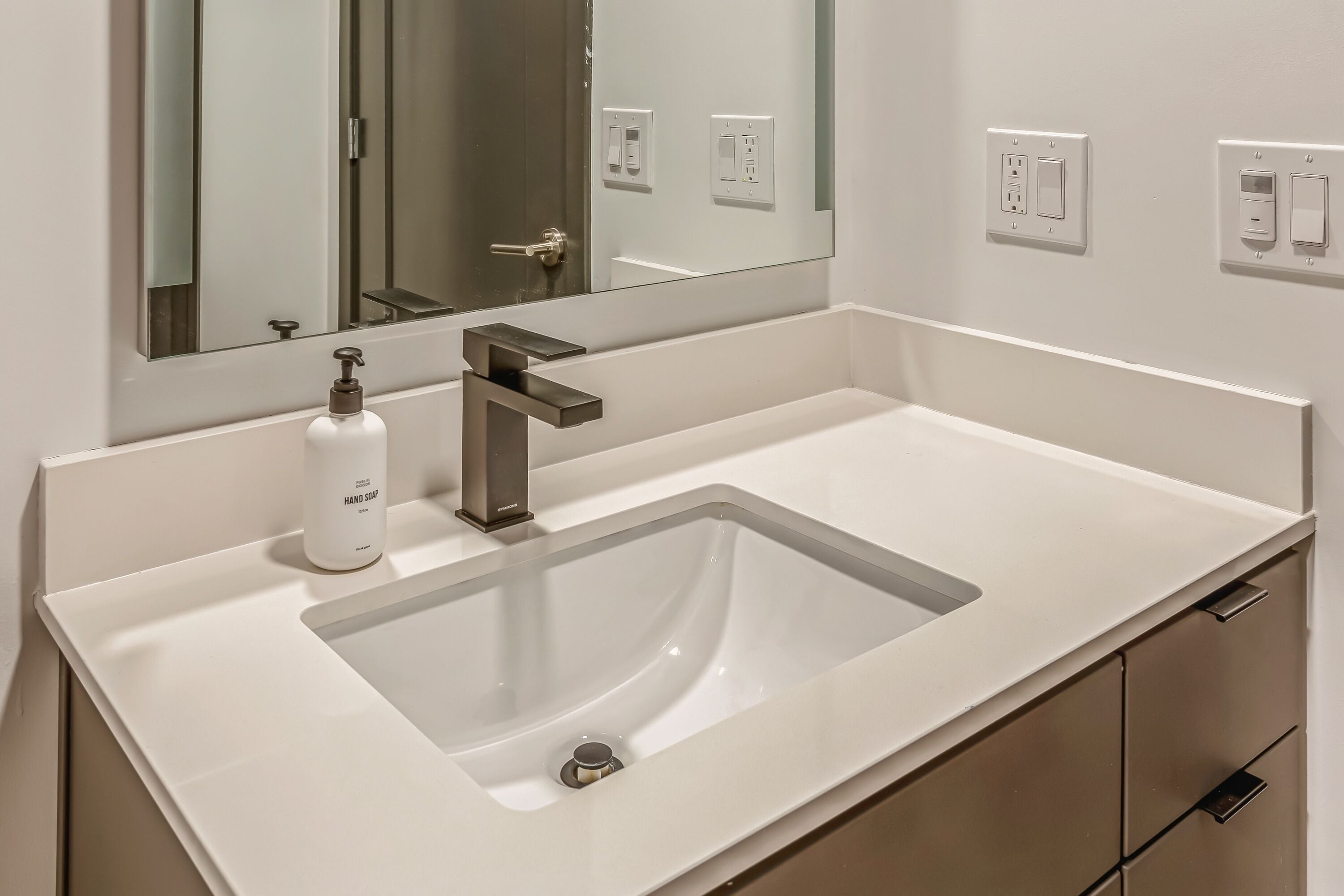 A modern bathroom with a white sink, a large mirror, and a bottle of hand soap.