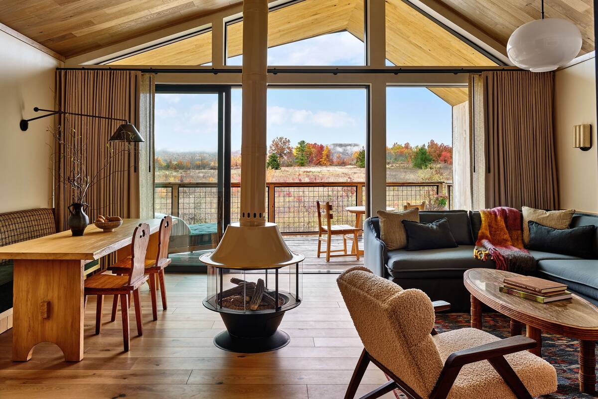 Ein geräumiges Wohnzimmer mit hoher Decke, großen Fenstern, einem Kamin und Blick auf einen Wald.