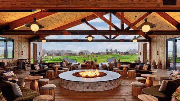 Amplia zona de estar al aire libre con una chimenea central, suelo de madera y vistas a un paisaje verde.