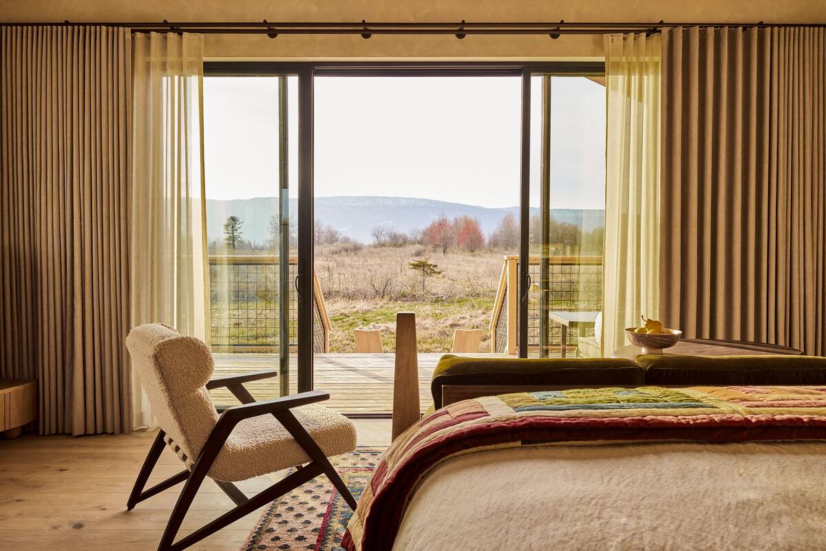Ein Schlafzimmer mit einem großen Bett, einem Sessel und einem Tisch mit Obstschale. Das Zimmer bietet einen Blick auf eine Landschaft mit Bäumen und Hügeln.