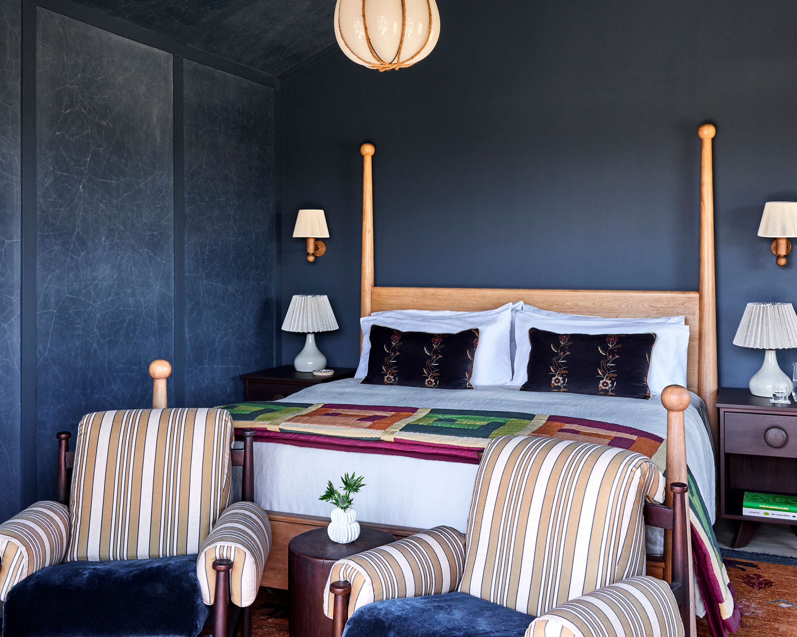 Un dormitorio con una cama grande, dos mesitas de noche, dos lámparas, un cabecero de madera, sillas a rayas y una pequeña mesa con un florero.