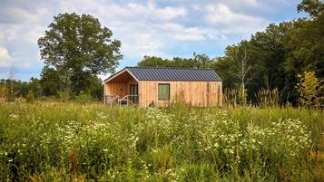 A wooden cabin with a metal roof, surrounded by tall grass and wildflowers, with trees in the background.