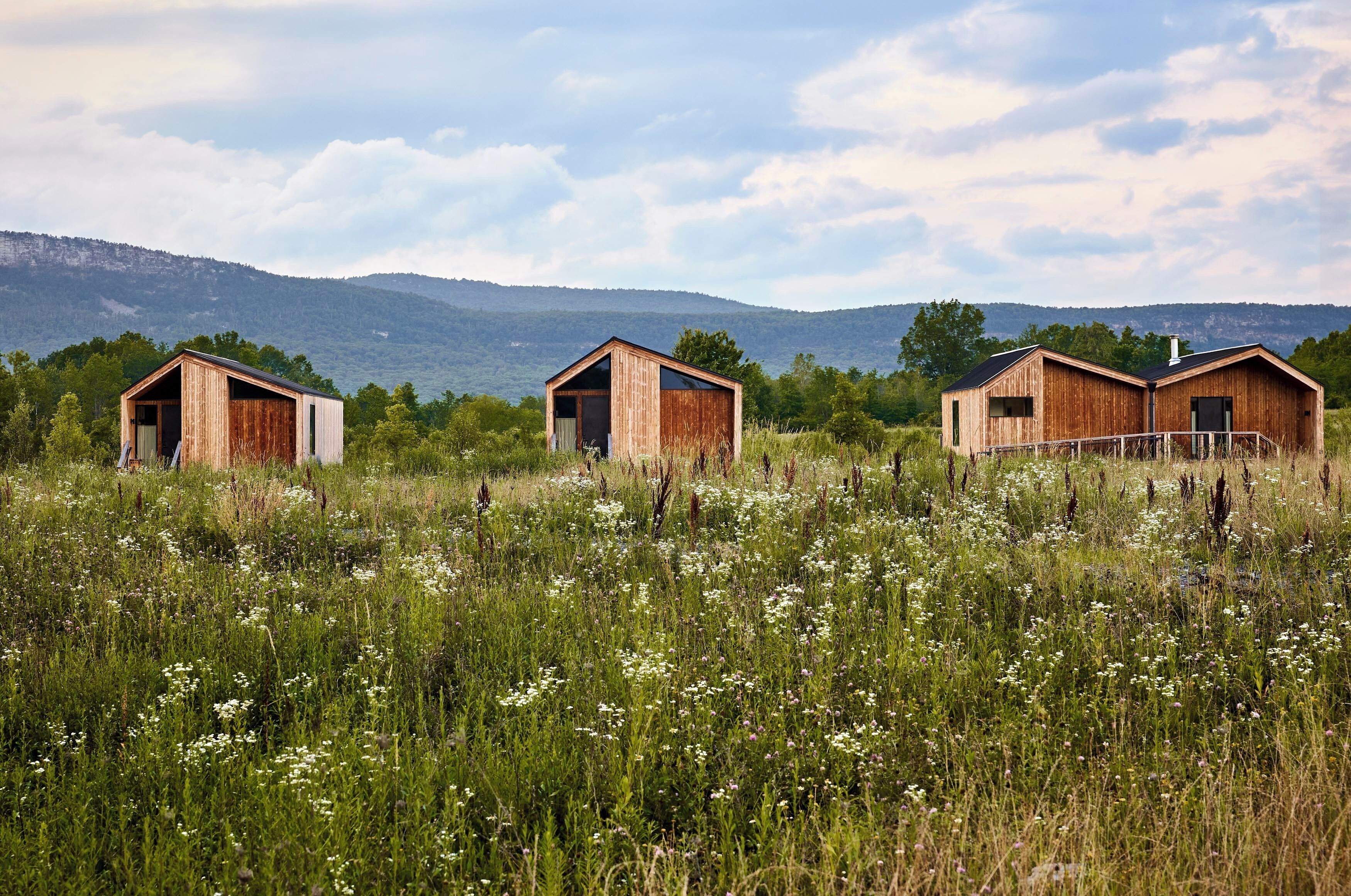 Three rustic wooden cabins in a grassy field with a mountainous backdrop.