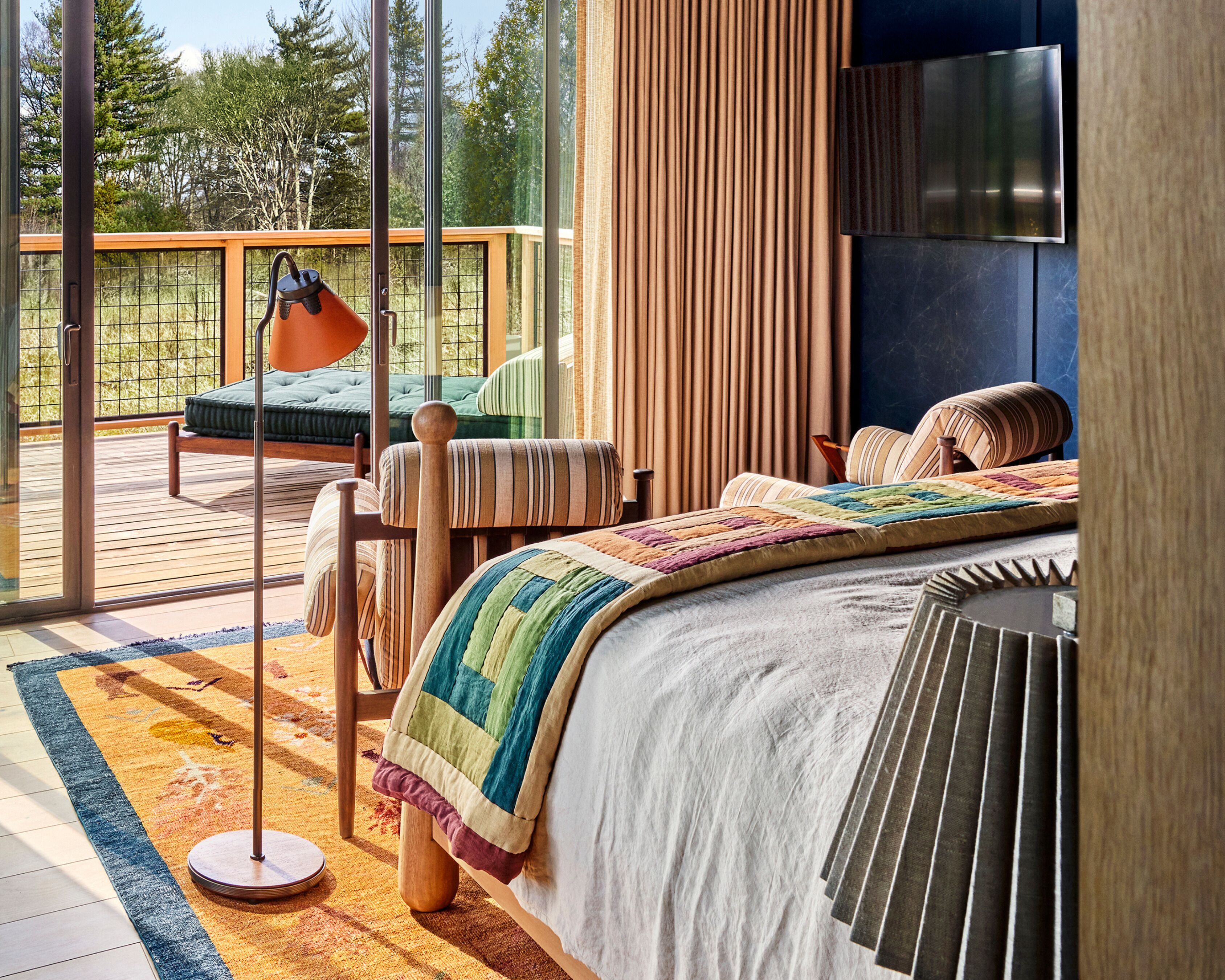 A bedroom with a large window, a bed with a colorful quilt, a wooden floor, and a view of the outdoors.