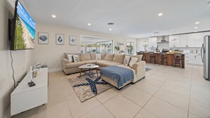 Ein modernes Wohnzimmer mit einem beigen Sofa, einem Couchtisch und einem Teppich mit Seestern-Muster. Im Hintergrund ist eine Küche mit weißen Schränken und einem Kühlschrank zu sehen.