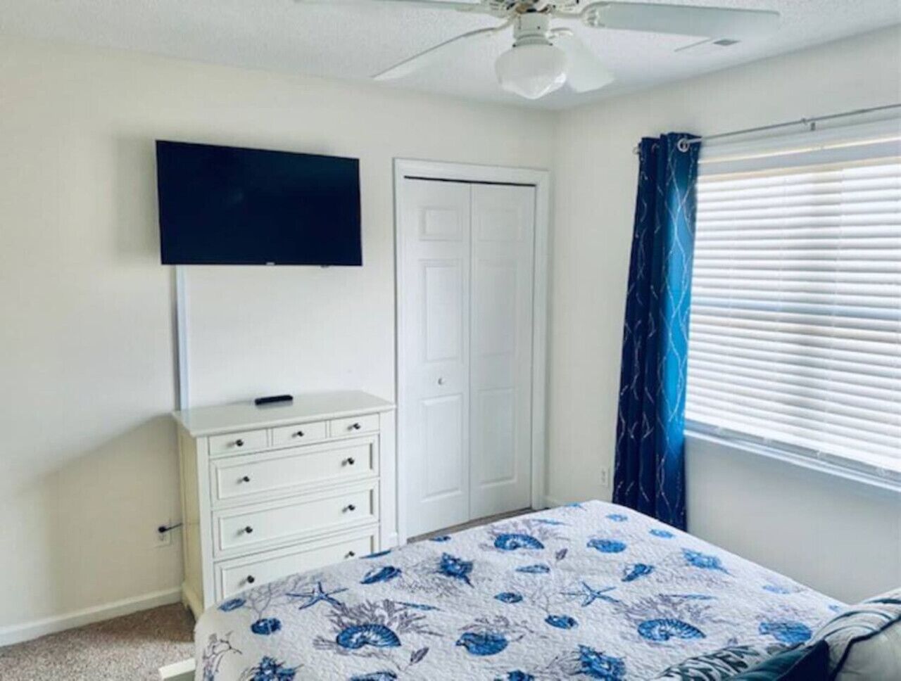 Een slaapkamer met een bed, een dressoir, een aan de muur bevestigde televisie, een deur en een raam met jaloezieën.