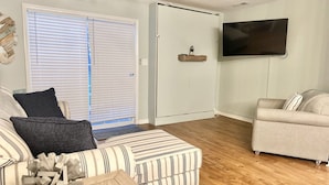 Un salon avec un canapé, un pouf et un téléviseur fixé au mur.