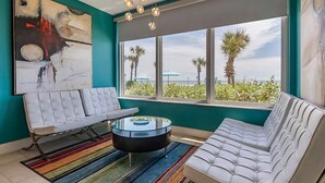 A modern living room with a white sofa, a white armchair, and a glass coffee table. There is a large window with a view of palm trees and the ocean. - Beautiful Daytona Beach Oceanfront 2 Bedroom Condo (Daytona Beach)