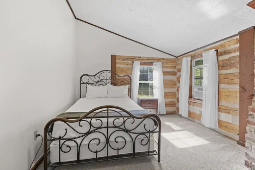 Un dormitorio con estructura de cama metálica, ropa de cama blanca, pared de madera y una ventana con cortinas.