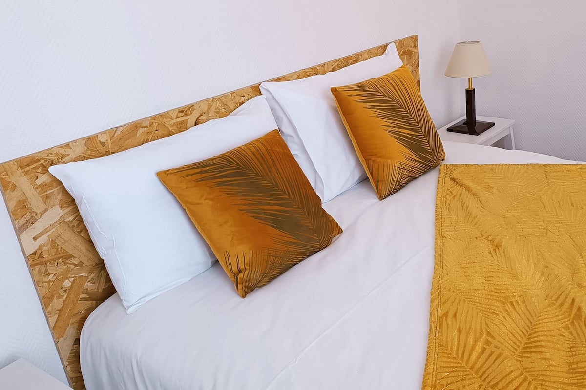 Un lit bien fait, avec une literie blanche et jaune moutarde, une tête de lit en bois et une lampe de chevet.