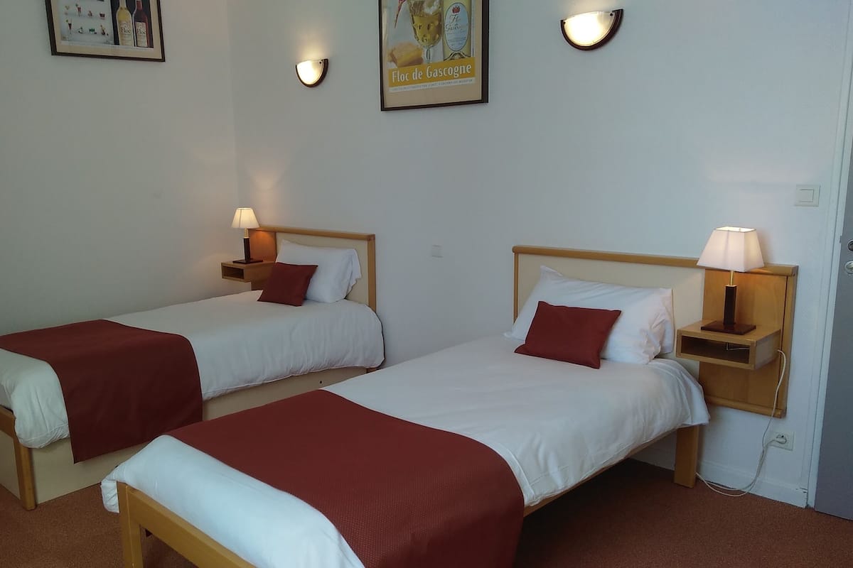 Une chambre d’hôtel avec deux lits simples, chacun doté d’une tête de lit en bois, de draps blancs et d’un plaid rouge. On y trouve des tables de chevet avec des lampes, une affiche encadrée au mur et une porte sur la droite.