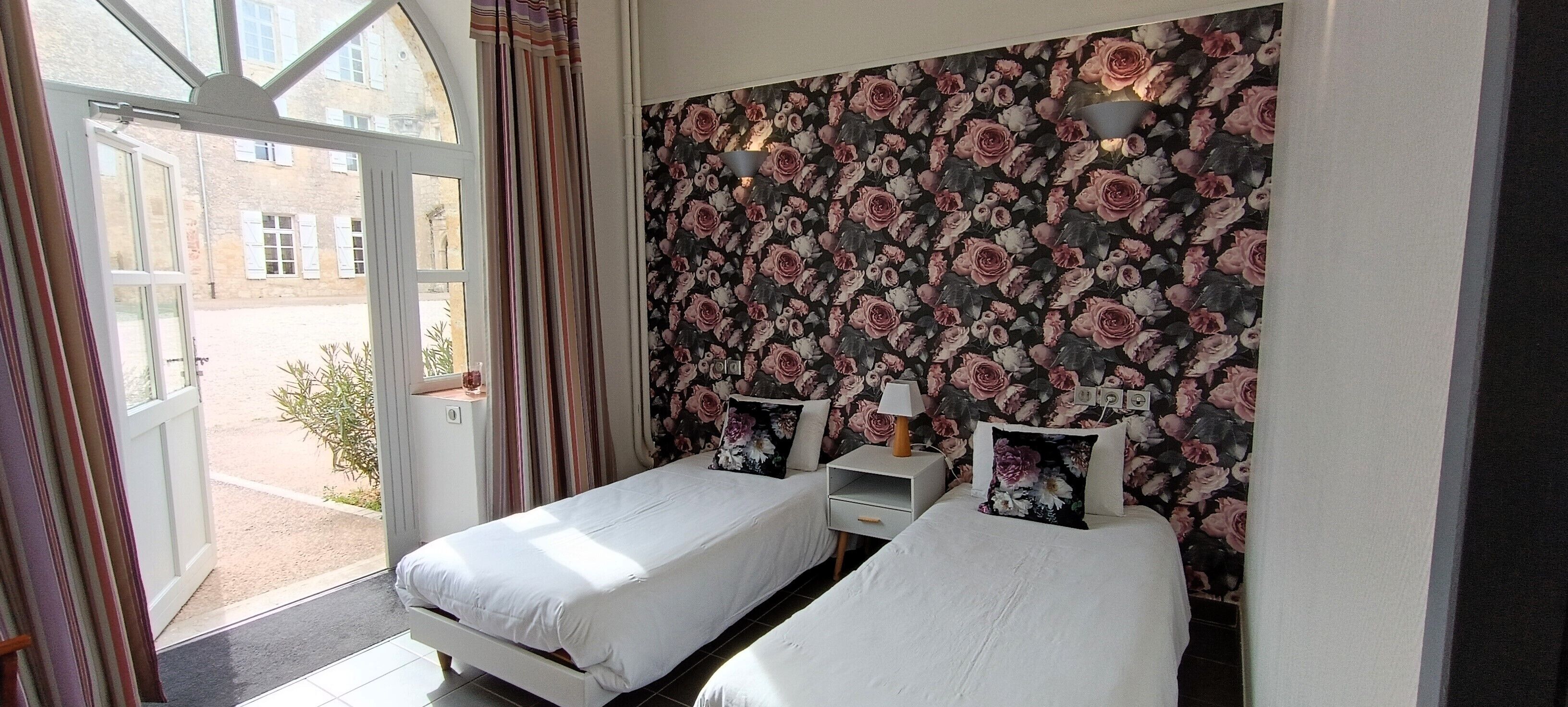 A bedroom with two single beds, a floral wallpaper, a window with curtains, and a small bedside table with a lamp.