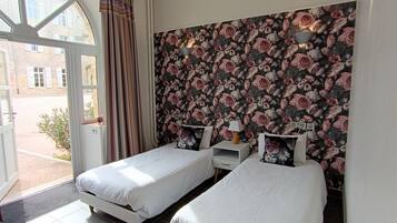 A bedroom with two single beds, a floral wallpaper, a window with curtains, and a small bedside table with a lamp.