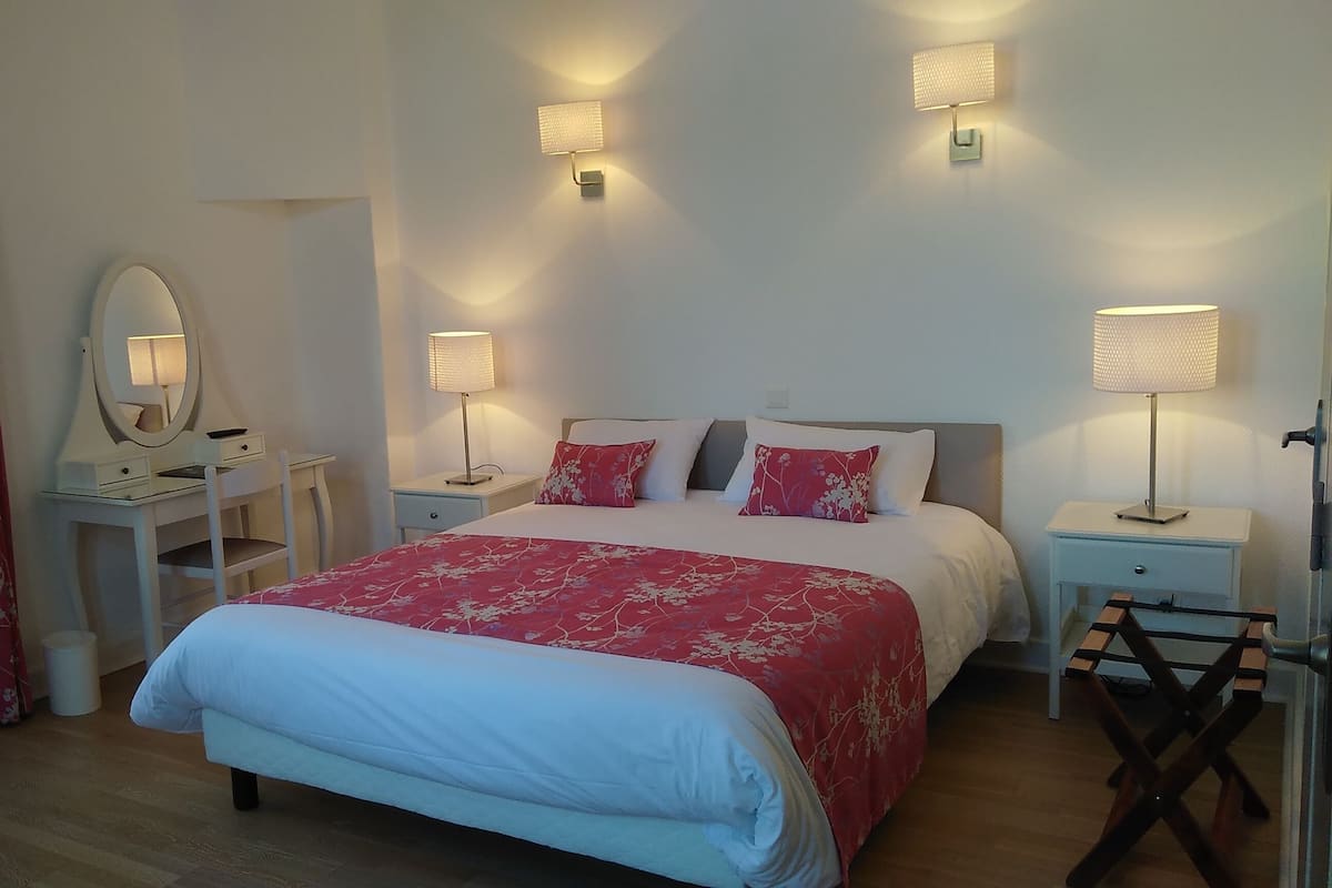 Une chambre à coucher avec un lit, des tables de chevet, une commode, un miroir et une fenêtre avec des rideaux.
