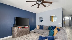 Ein Wohnzimmer mit Flachbildfernseher, Deckenventilator, einer Couch mit Kissen und einem gemusterten Teppich.