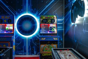 A retro arcade with multiple gaming machines, a central neon ring, and a cityscape view through the window. - Walk to Clubhouse | Tron Game Room | Tiki Bar | Private Pool | 7BR Villa (Kissimmee)