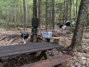 Una mesa de picnic de madera en un bosque con una parrilla y un generador portátil.