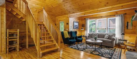 A wooden cabin interior with a staircase, a living room with a sofa, chairs, and a coffee table.
