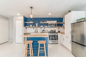 A modern kitchen with a blue accent wall, wooden bar stools, stainless steel appliances, and a hanging light fixture. - A SA Pearl (San Antonio)