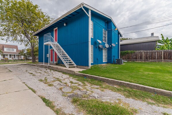 A blue building with a red door and white steps, surrounded by a grassy yard and a gravel path. - A SA Pearl (San Antonio)